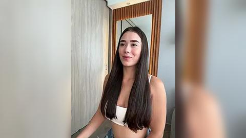 Media: Video of a young woman with long, straight black hair, wearing a white bra, standing in a modern, minimalist room with light gray walls and wooden accents.