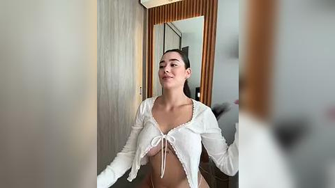 Media: Video of a young woman with fair skin and long dark hair, wearing a white, ruffled crop top, standing in a modern room with wooden and glass accents.