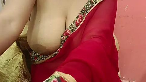 Media: Video of a woman with light skin, wearing a red saree with gold floral embroidery. Her left breast is partially exposed, showing a natural breast shape. Background shows a pink wall and a beige blanket.