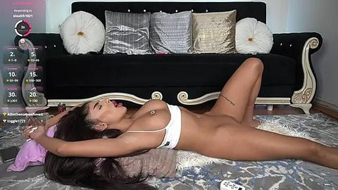 Media: A video shows a nude woman with dark skin, long dark hair, and large breasts, lying on a patterned rug in a room with a black sofa adorned with silver and white cushions.