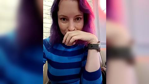 Media: Video of a woman with shoulder-length purple hair, wearing a blue and black striped sweater, sitting indoors, hands clasped under chin, blurred background.