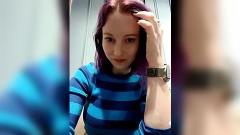 Media: Video of a young woman with purple hair, wearing a blue-striped shirt, resting her hand on her head, in a blurry background.