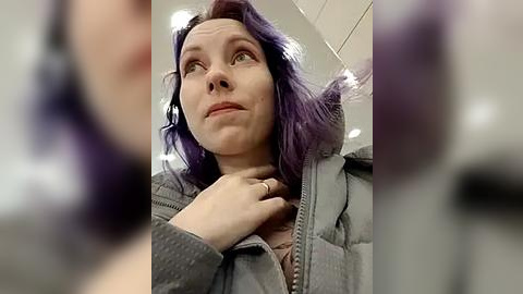 Media: A video of a woman with shoulder-length, wavy purple hair, wearing a gray puffer jacket, touching her collar, looking contemplative in a dimly lit indoor setting.