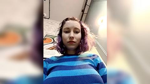 Media: Video of a young woman with shoulder-length, wavy, purple hair, wearing a blue and black striped sweater, standing in a brightly lit kitchen with white brick walls and a visible recipe book.