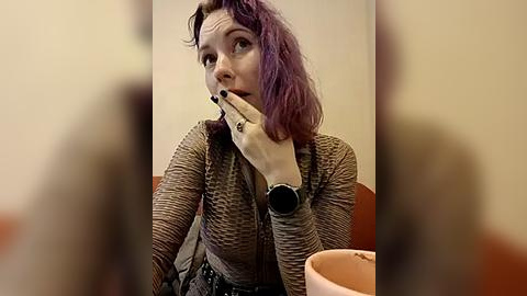 Media: Video of a fair-skinned woman with shoulder-length purple hair, wearing a brown cardigan, black nail polish, and a black watch, sitting at a restaurant table with an orange cup, looking thoughtful.