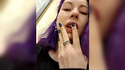 Media: Video of a young woman with vivid purple hair, light skin, and blue nail polish, licking her fingers while wearing multiple rings. Background shows a blurred indoor setting with beige walls and a grid window.