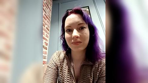 Media: Video of a fair-skinned woman with long, wavy, purple hair, wearing a textured, brown cardigan. Background features a brick wall and a blue door, creating a cozy, indoor setting.