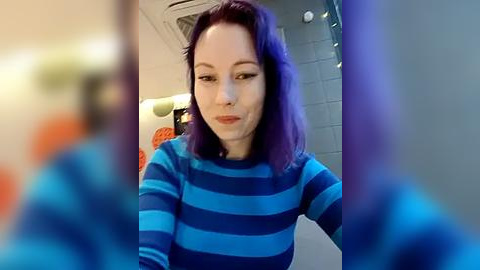 Media: Video of a young woman with shoulder-length purple hair, wearing a blue and white striped sweater, smiling in a bathroom with tiled walls and a blurred orange object in the foreground.