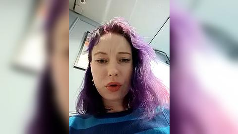 Media: A video of a woman with shoulder-length, vibrant purple hair, wearing a blue striped sweater, taken from a low angle, with a blurry background.