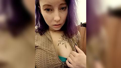 Media: Video of a young woman with purple hair, fair skin, and a tattoo of a leaf on her chest, wearing a brown knit sweater, and holding a teal bra.