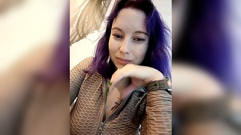 Media: A video of a young woman with purple hair, fair skin, and wearing a textured brown top, sitting with her elbow on a table, her hand supporting her chin, in a dimly lit room.