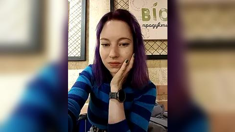 Media: Video of a woman with purple hair, wearing a blue striped shirt, leaning on her hand, in a cafe with \"Biogems\" logo on the wall.