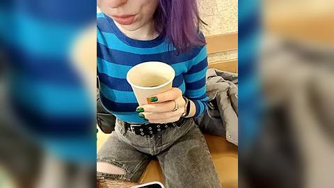 Media: Video of a young woman with purple hair, wearing a blue and black striped shirt, drinking coffee, sitting on a wooden floor, and wearing ripped jeans.