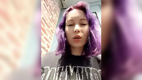 Media: A video of a young woman with purple hair, wearing a black graphic t-shirt, standing in front of a brick wall, slightly out of focus.