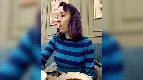 Media: Video of a woman with purple hair, wearing a blue and black striped top, sitting in a cozy, dimly lit cafe. The background features two framed posters and a teal cushioned chair.