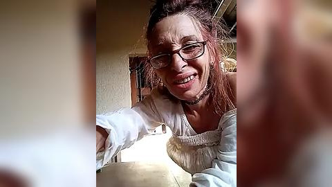 Media: Video of a middle-aged woman with glasses and messy hair, wearing a white blouse, laughing while holding a beige bowl, in a dimly lit kitchen.