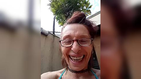 Media: A video of a Caucasian woman with red hair, glasses, and a black choker, laughing, standing outdoors with a concrete wall and greenery in the background.