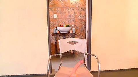 Media: Video of a small, cozy bathroom with a cream-colored wall and tiled backsplash. A white chair with a male person's erect penis visible, facing a sink with toiletries and a mirror.