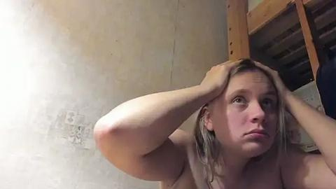 Media: Video of a topless, fair-skinned woman with shoulder-length blonde hair, looking up with hands on her head, in a dimly lit room with a wooden bunk bed and peeling wallpaper.