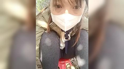 Media: A video captures a young woman in a green jacket, blue jeans, and a white face mask, kneeling on the ground with her hands tied behind her back. The background features blurry figures and foliage.