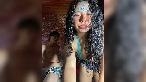 Media: Video: A young, dark-skinned woman with curly hair and blue face paint, wearing a blue bikini, stands in front of a brick wall. Behind her, a shirtless, light-skinned man sits on a bed, wearing only striped underwear.