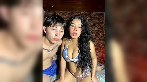 Media: Video of a young, attractive couple in a dimly lit room. The man, with short brown hair, wears blue shorts; the woman, with long black hair, wears a revealing white and blue bikini. Brick wall background.