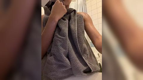 Media: Video of a person with light brown skin, wrapped in a gray towel, standing in a bathroom with white tiled walls and a shower curtain. The image is slightly blurred, with the focus on the towel.