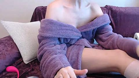 Media: A video of a fair-skinned person in a purple, off-the-shoulder robe, sitting on a plush, dark purple couch, with a white textured pillow and a pink vibrator nearby.