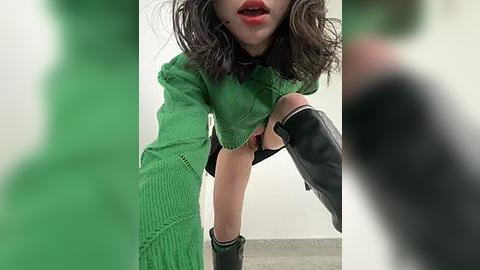 Media: Video of a woman in a green, textured sweater, black skirt, and knee-high boots, bending over in an indoor setting with blurred green objects in the foreground.