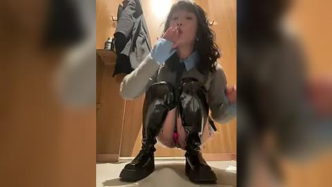 Media: Video of a young Asian woman squatting on a carpeted floor, wearing a gray sweater, black leather mini-skirt, thigh-high boots, and pink panties. She has long black hair and is holding a stuffed animal.