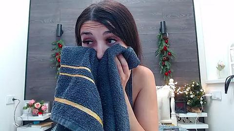 Media: Video of a young woman with long brown hair, covering her face with a dark blue towel, in a modern, well-lit bathroom with festive holiday decorations.