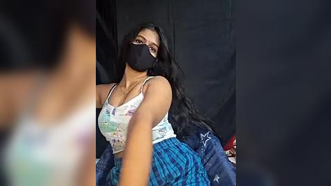 Media: Video of a young woman with long black hair, wearing a black face mask, a white tank top with a colorful floral design, and blue plaid skirt, seated against a dark curtain background.