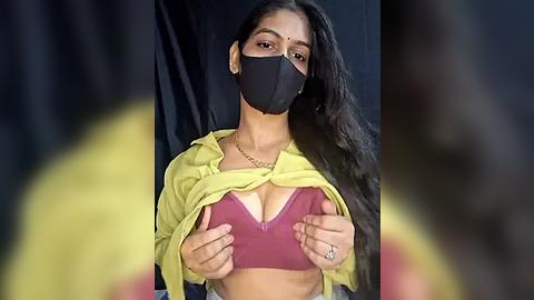 Media: A video of a young woman with long black hair, wearing a yellow hoodie, a black mask, and a maroon bra, lifting her hoodie to reveal her cleavage, against a dark backdrop.