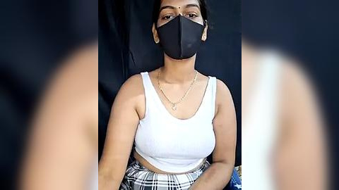 Media: Video of a young woman with medium brown skin, wearing a black face mask, white tank top, and plaid skirt, seated against a black backdrop.