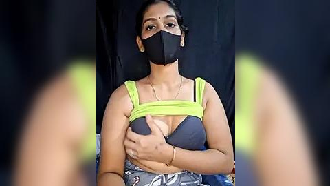 Media: Video of a young South Asian woman with medium skin tone, wearing a neon green crop top, black face mask, and floral-patterned skirt, seated against a black backdrop.