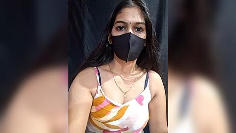 Media: Video of a South Asian woman with medium brown skin, long dark hair, wearing a black face mask, white and pink floral-patterned top, and gold necklace.