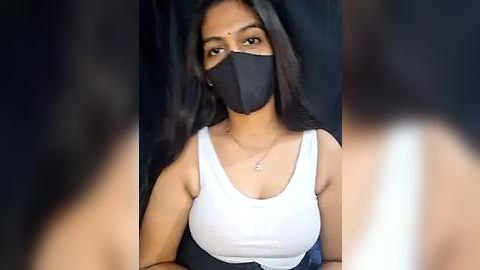 Media: Video of a young woman with medium skin tone, long black hair, wearing a white tank top, black mask, and blue denim shorts, standing against a blurred dark background.