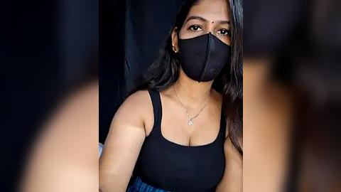 Media: Video of a South Asian woman with medium skin tone and long black hair, wearing a black face mask, black tank top, and blue skirt, seated indoors against a dark backdrop.