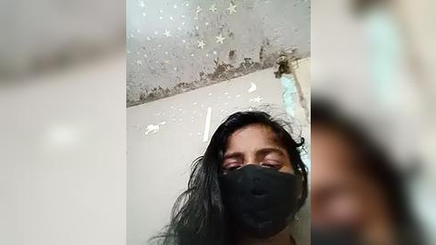 Media: A video of a woman with long black hair and a black mask, standing in a room with peeling white walls and a ceiling dotted with stars. The image is slightly blurry.