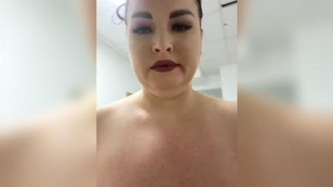 Media: A close-up video of a fair-skinned, plus-sized woman with dark eyebrows, wearing dark lipstick, and a nose ring. Her face is partially blurred, emphasizing her full cheeks and round features. The background is an indoor setting with white walls and a ceiling.