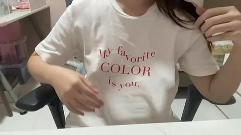 Media: Video of a woman in a white T-shirt with red text: \"My favorite color is you.\" She sits at a table, left arm resting on it, with a pink bag and a counter in the background.