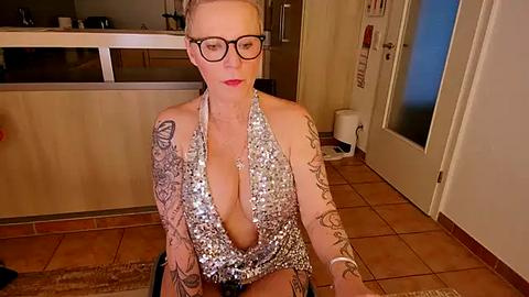 Media: A video of a blonde woman with tattoos, wearing a silver sequin halter top and black glasses, sitting on a tiled floor in a kitchen.