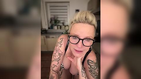 Media: Video of a middle-aged blonde woman with glasses, tattoos, and black lingerie, leaning against a modern kitchen counter, smiling and blowing a kiss.