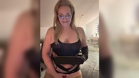 Media: Video of a blonde woman with glasses, wearing a black fishnet top, black gloves, and black panties, holding a tablet in a dimly-lit bedroom.