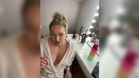 Media: Video of a middle-aged woman with glasses, blonde hair in a bun, wearing a white robe, sitting at a vanity in a dimly lit room with makeup brushes and bottles.