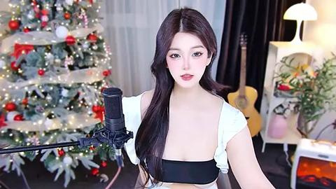 Media: Video of an East Asian woman with long black hair, wearing a black bra and white top, singing into a microphone in a festive, decorated living room with a Christmas tree and guitar.