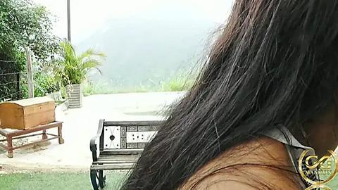 Media: Video of a person's shoulder-length brown hair, outdoor patio with white bench, wooden table, green plants, and a foggy mountainous background.