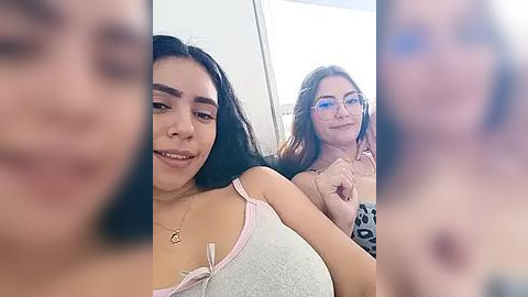 Media: Video of two smiling women, one with long black hair and glasses, wearing a white tank top, and the other with long brown hair, also in glasses, both indoors.