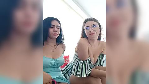 Media: Video of two women seated, one with long dark hair, wearing a blue top, and the other with glasses and a striped off-shoulder top.