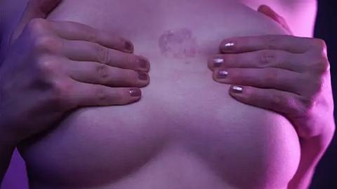 Media: Video of a light-skinned person's bare chest, with small breasts, hands holding them, and metallic nail polish, against a purple-lit background.